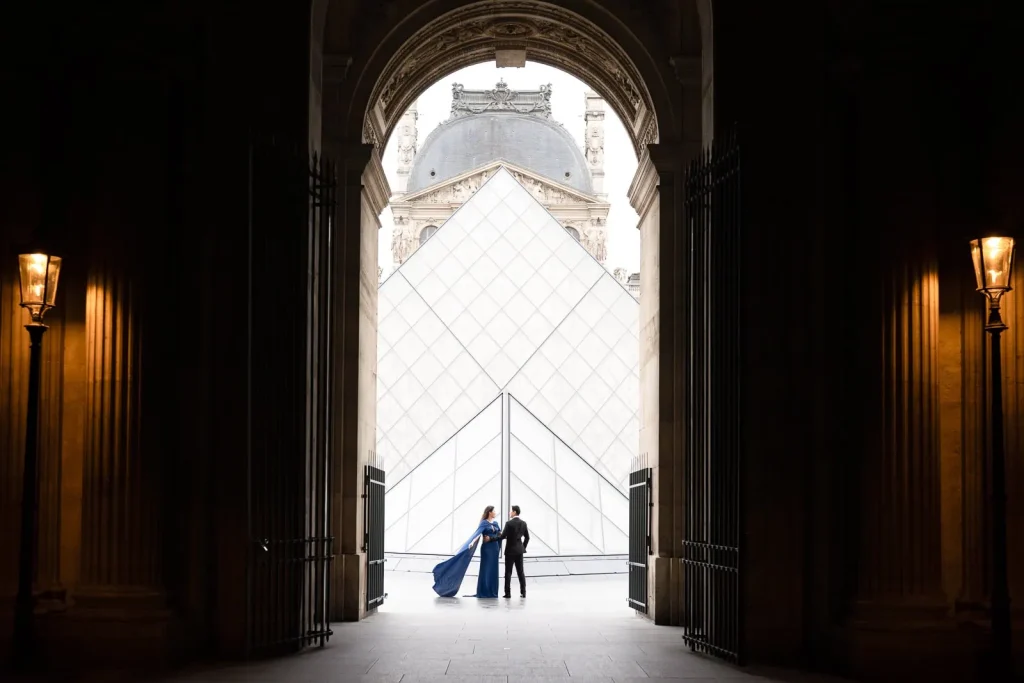Anniversary couple in blue gown and black suit posing at Louvre Pyramid from Cour Napoleon underpass with warm lantern lighting at dusk