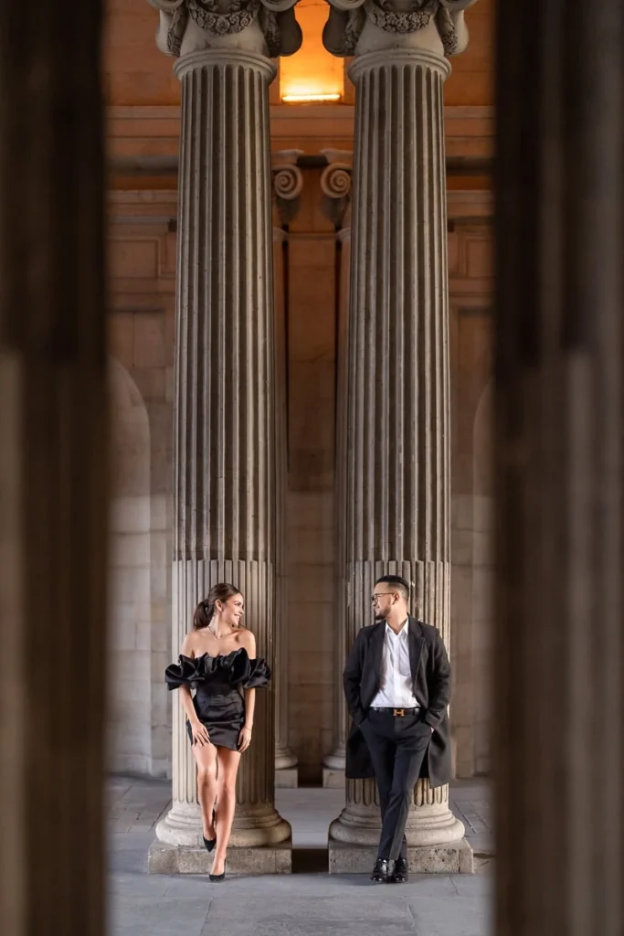 Stylish couple posing on either side of Louvre underpass columns at golden hour, her silhouette lit by warm sunlight
