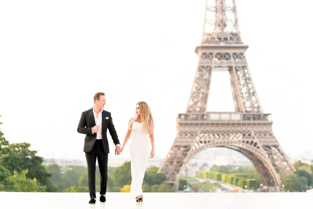 A stylish couple wearing Louboutin shoes holds hands in a walking pose at sunrise on an empty Trocadéro platform, with the Eiffel Tower in the background.