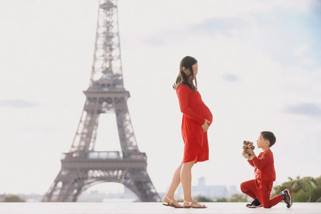 Young boy on knee presenting teddy bear to pregnant mother at Trocadéro sunrise with Eiffel Tower, Paris maternity photographer
